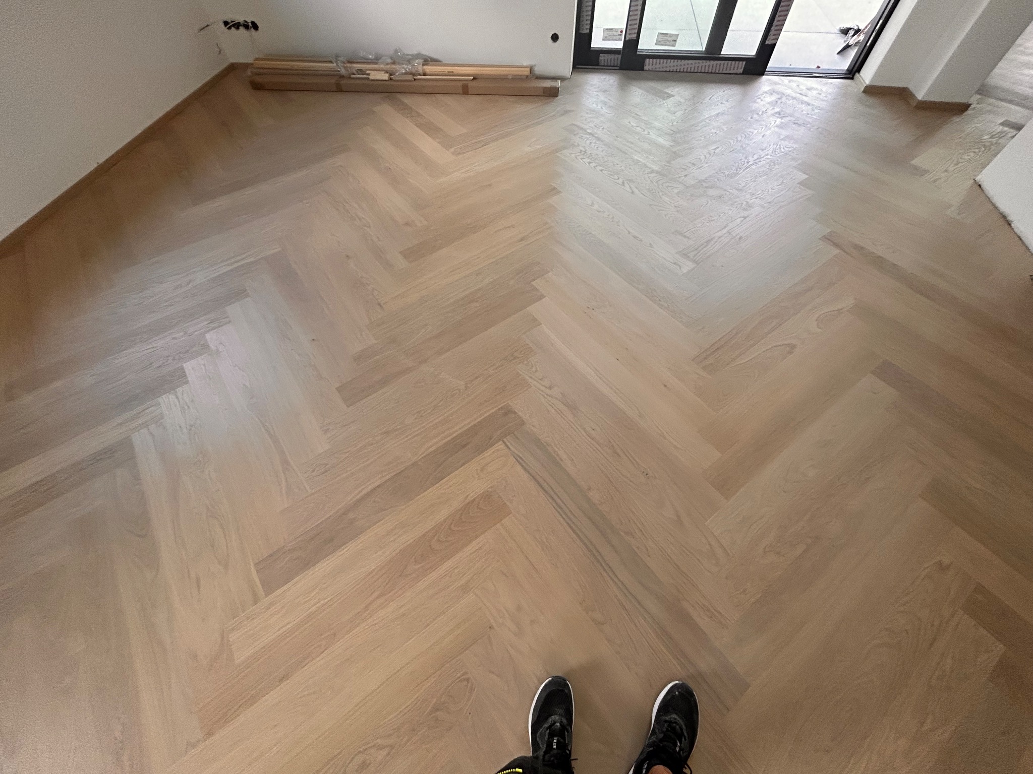 Jasny parkiet ułożony we wzór jodełki francuskiej, widok z góry na fragment podłogi z parą butów w dolnej części kadru, w tle deski do ułożenia.