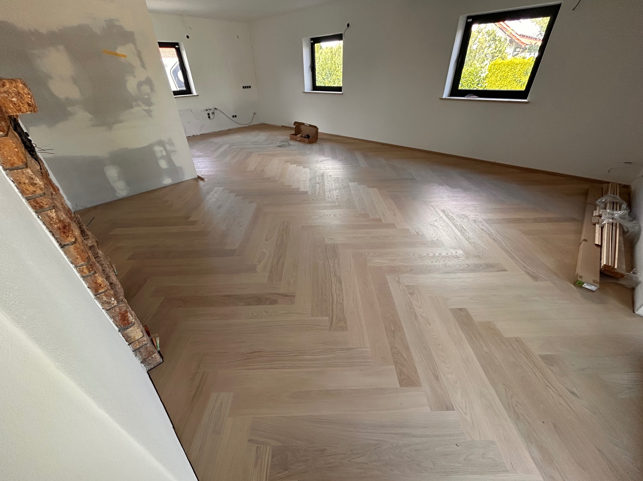 Jasny parkiet ułożony we wzór jodełki francuskiej w przestronnym, jasnym pokoju z trzema oknami. Fragment ściany z surowym wykończeniem i ceglanym elementem dekoracyjnym.