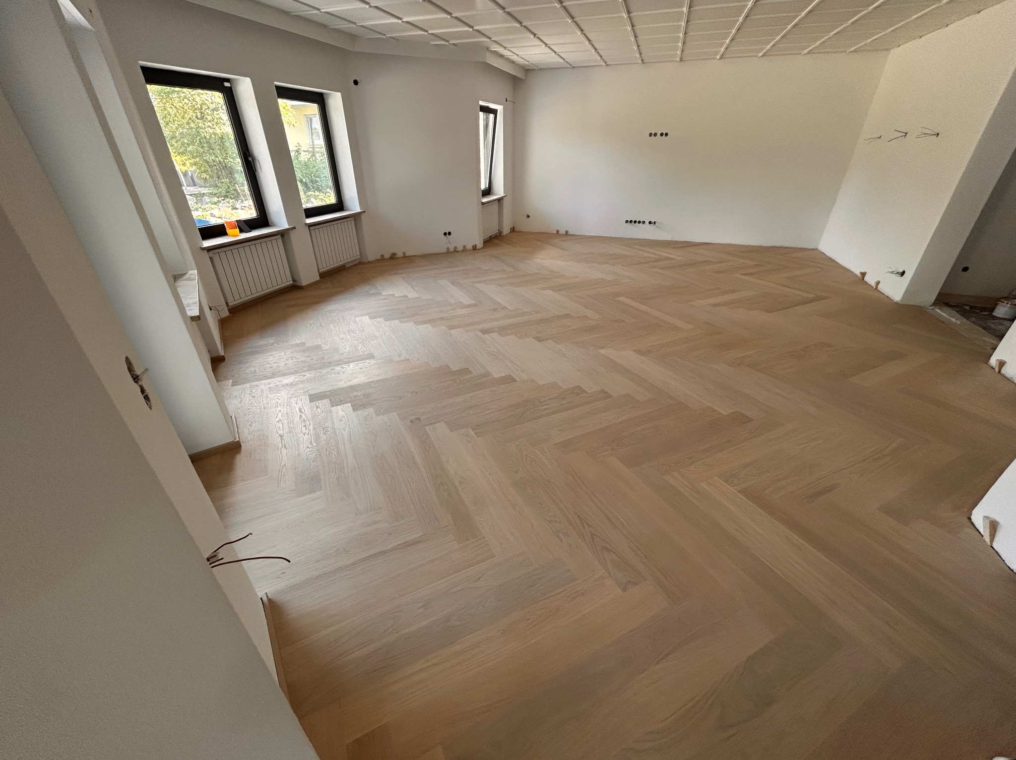 Jasny parkiet ułożony we wzór jodełki francuskiej w przestronnym, odnowionym pokoju z trzema oknami i dekoracyjnym sufitem kasetonowym. Widoczne niedokończone prace wykończeniowe.