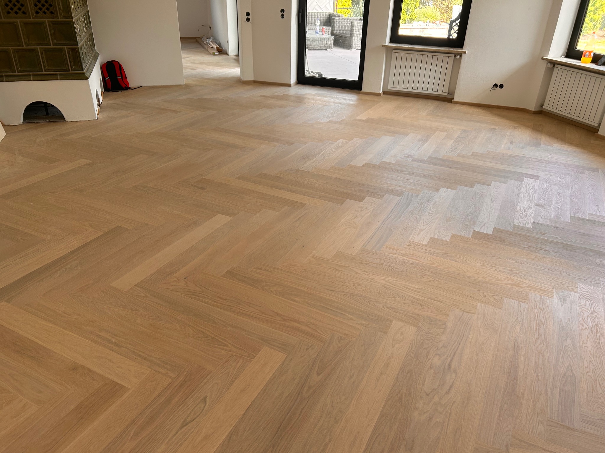 Jasny parkiet ułożony w jodełkę francuską w przestronnym wnętrzu z kominkiem kaflowym i dużymi oknami. Widoczne detale wykończenia i naturalne światło.