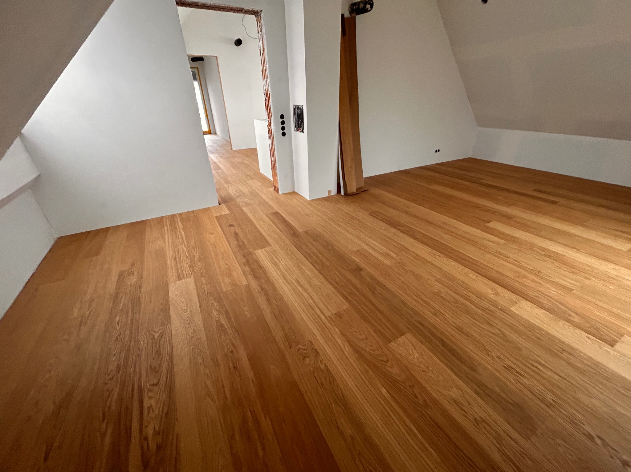 Jasny parkiet w nowym poddaszu, widoczne białe ściany i przygotowane otwory drzwiowe. Naturalne oświetlenie podkreśla ciepły odcień drewna.