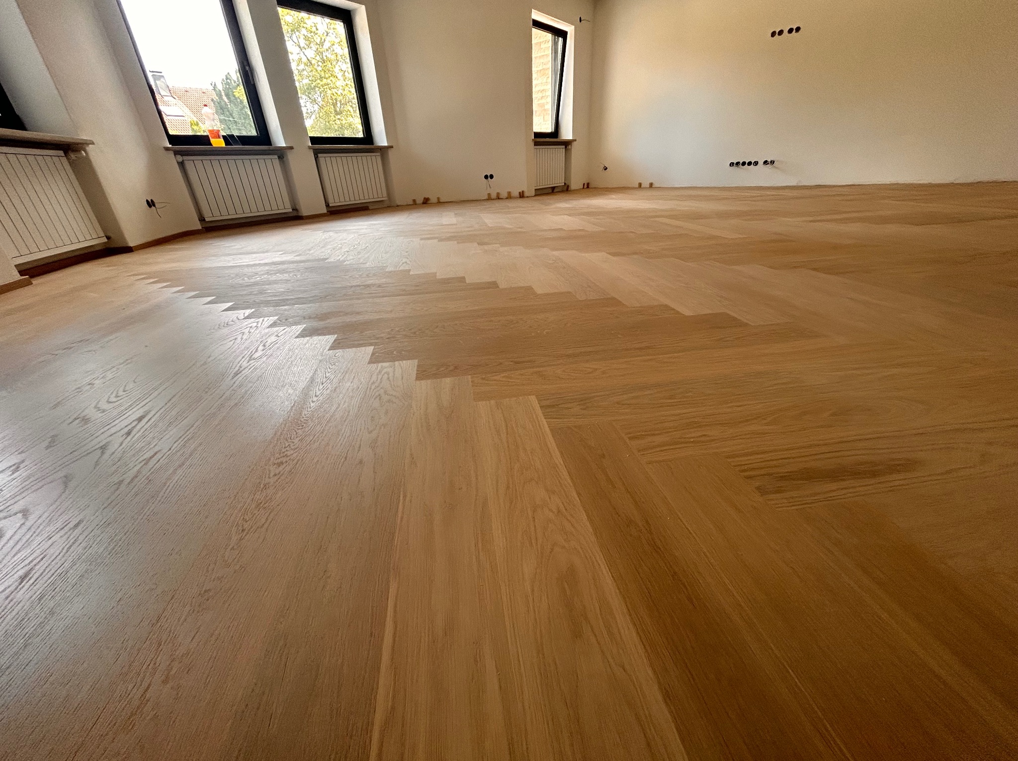 Jasny parkiet ułożony we wzór jodełki francuskiej w przestronnym, jasnym pokoju z oknami i grzejnikami. Widoczne listwy przypodłogowe i przygotowane punkty elektryczne na ścianie.