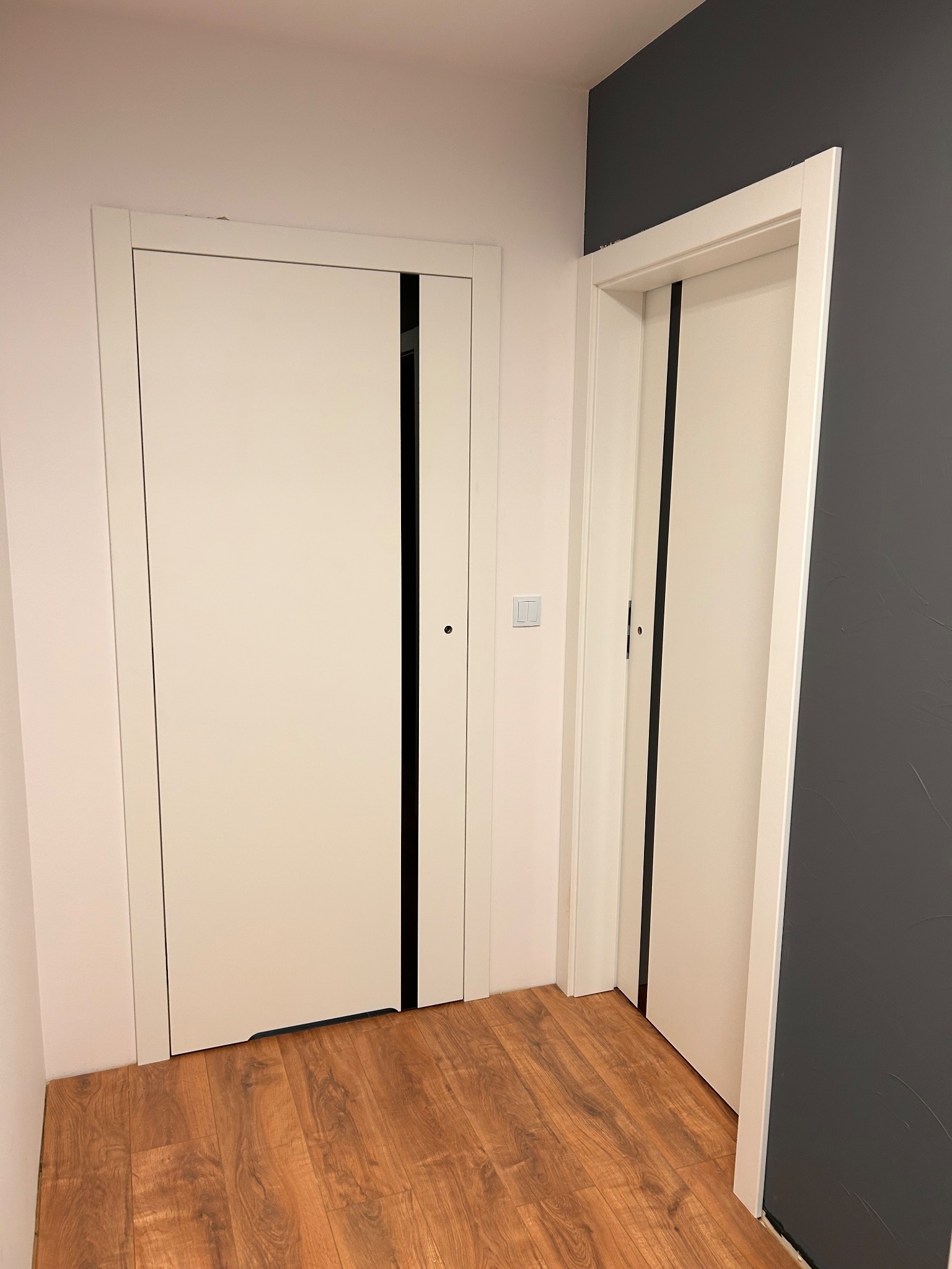 Dwa białe, nowoczesne drzwi wewnętrzne z czarnymi akcentami na tle drewnopodobnej podłogi i ściany w odcieniach bieli i grafitu. Widoczny fragment włącznika światła.