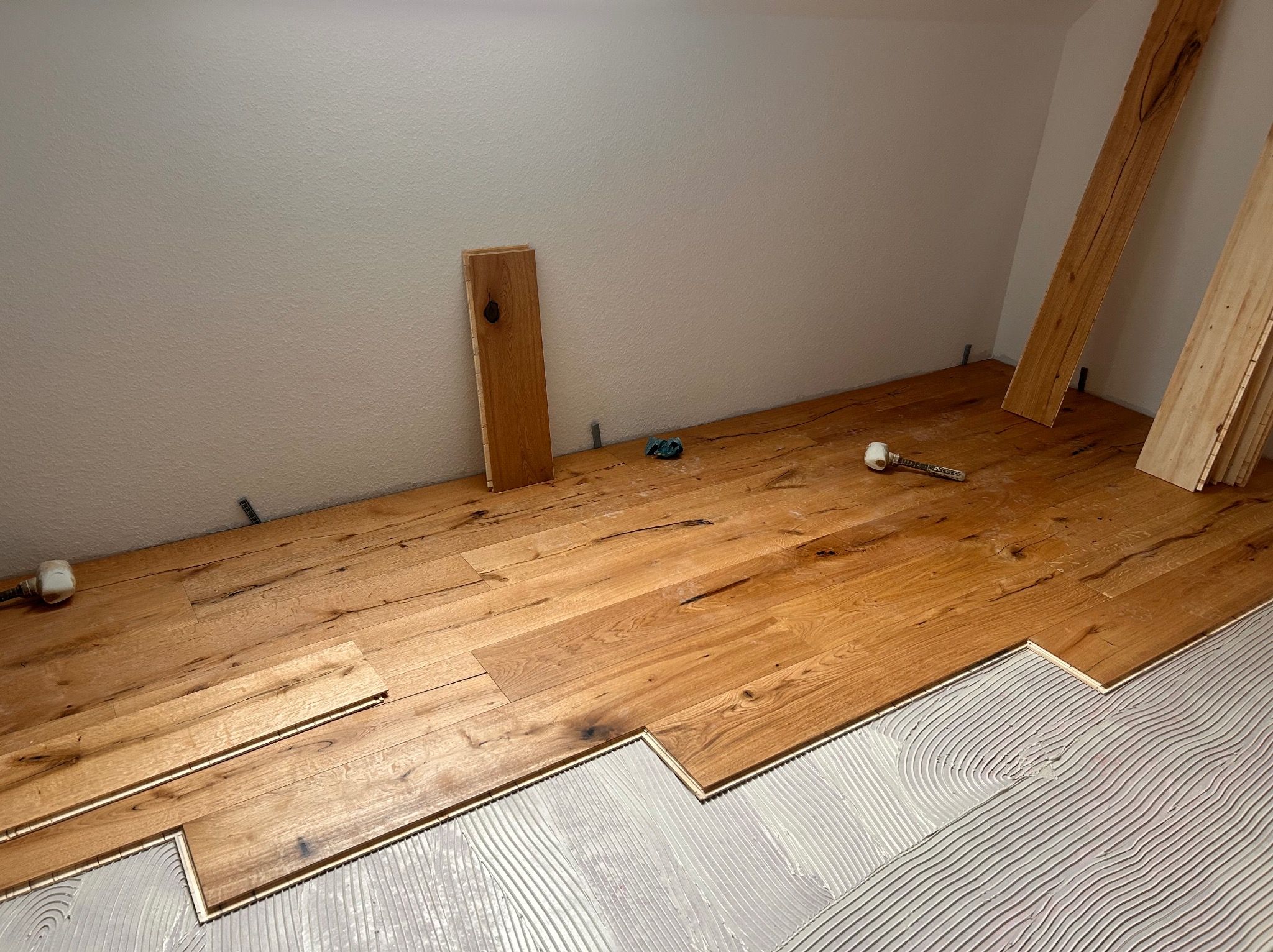 Układanie drewnianego parkietu na klej. Widoczne deski z naturalnego drewna i narzędzia. Częściowo ułożona podłoga w jasnym pomieszczeniu.