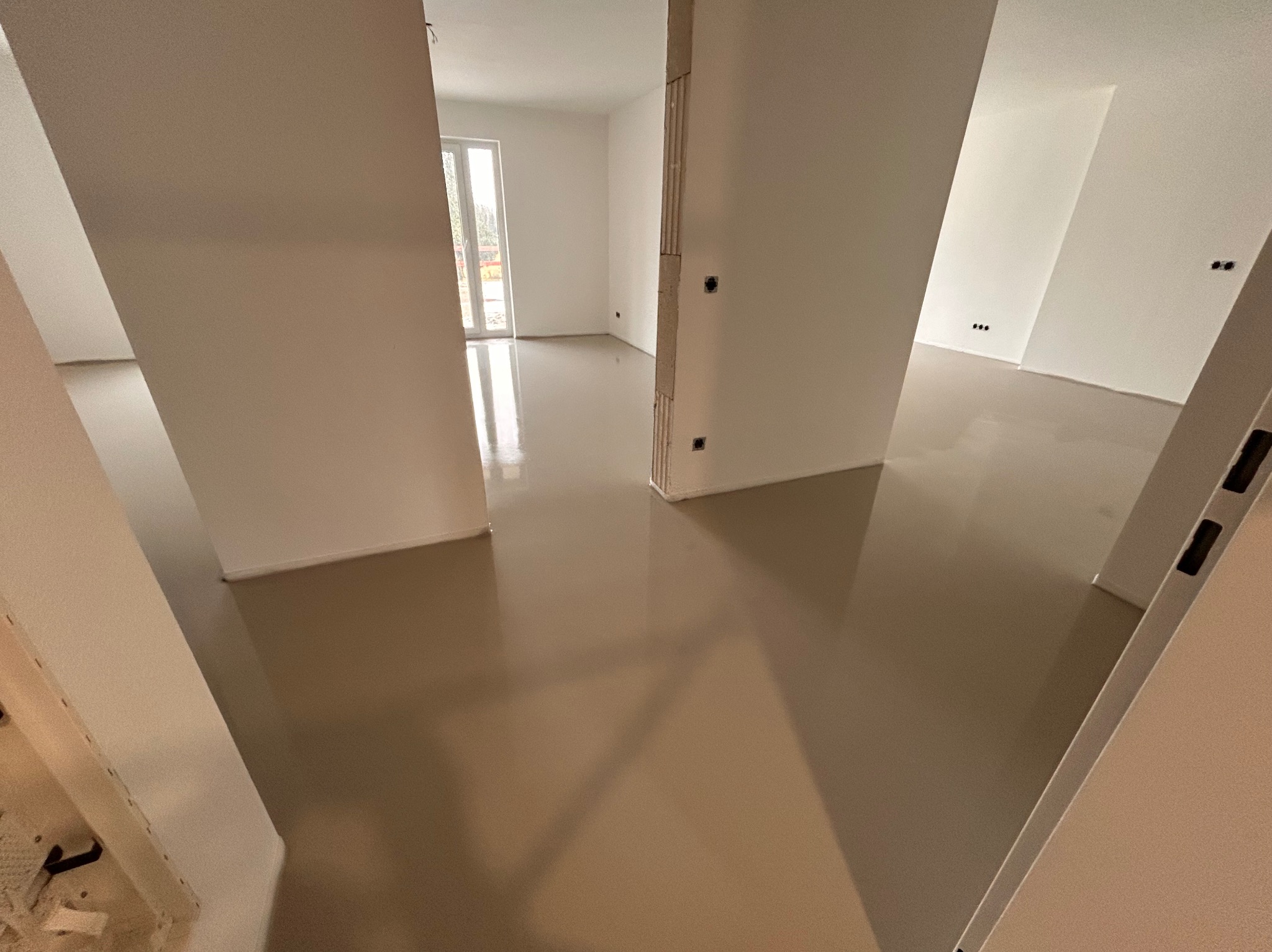 Świeżo wylana, beżowa posadzka w jasnym, pustym pomieszczeniu z widocznymi instalacjami elektrycznymi w ścianach. Gładka powierzchnia odbija światło.