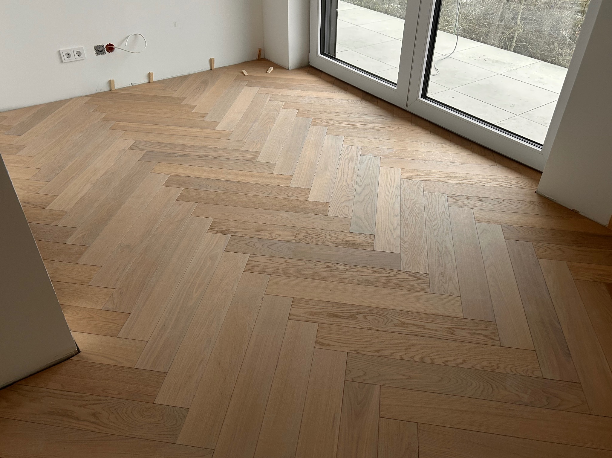 Jasny parkiet ułożony we wzór jodełki francuskiej w nowym mieszkaniu, widoczne gniazdka elektryczne i listwy przypodłogowe. Duże okno balkonowe w tle.