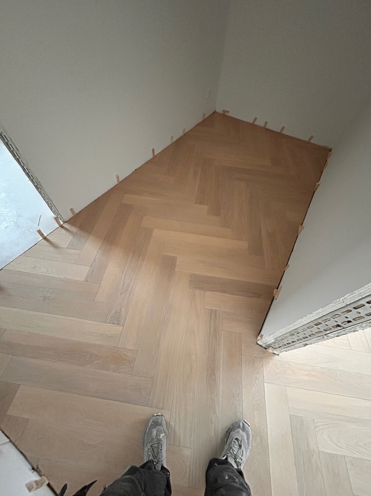 Jasny parkiet ułożony we wzór jodełki, z widocznymi klinami dystansowymi przy ścianach. U dołu kadru stopy w roboczych butach. Ujęcie z góry, w trakcie prac.