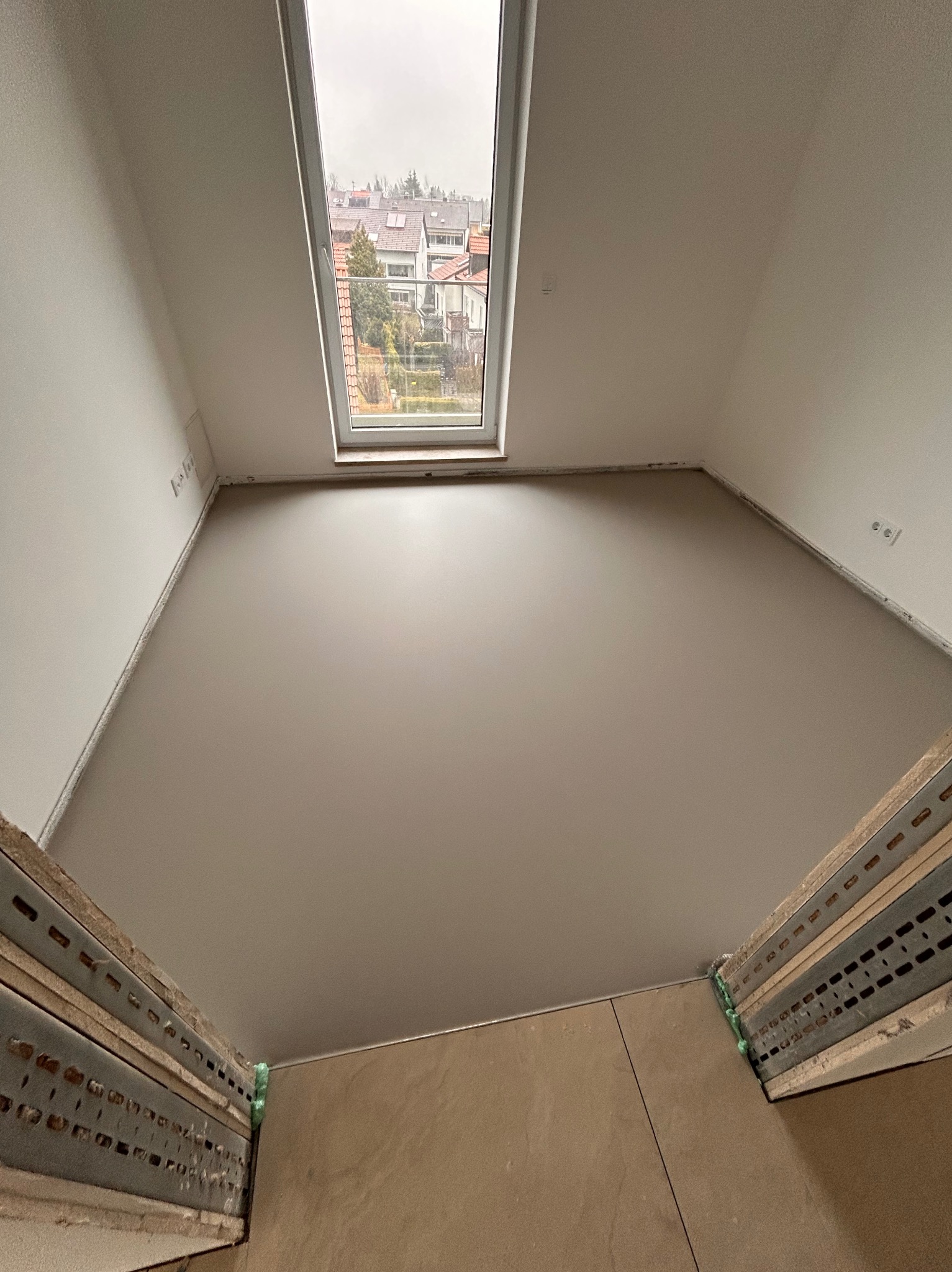 Poddasze po gładziowaniu ścian i sufitu, widok na okno dachowe i fragment podłogi. Metalowe profile w dolnej części kadru. Świeżo wykończone wnętrze.