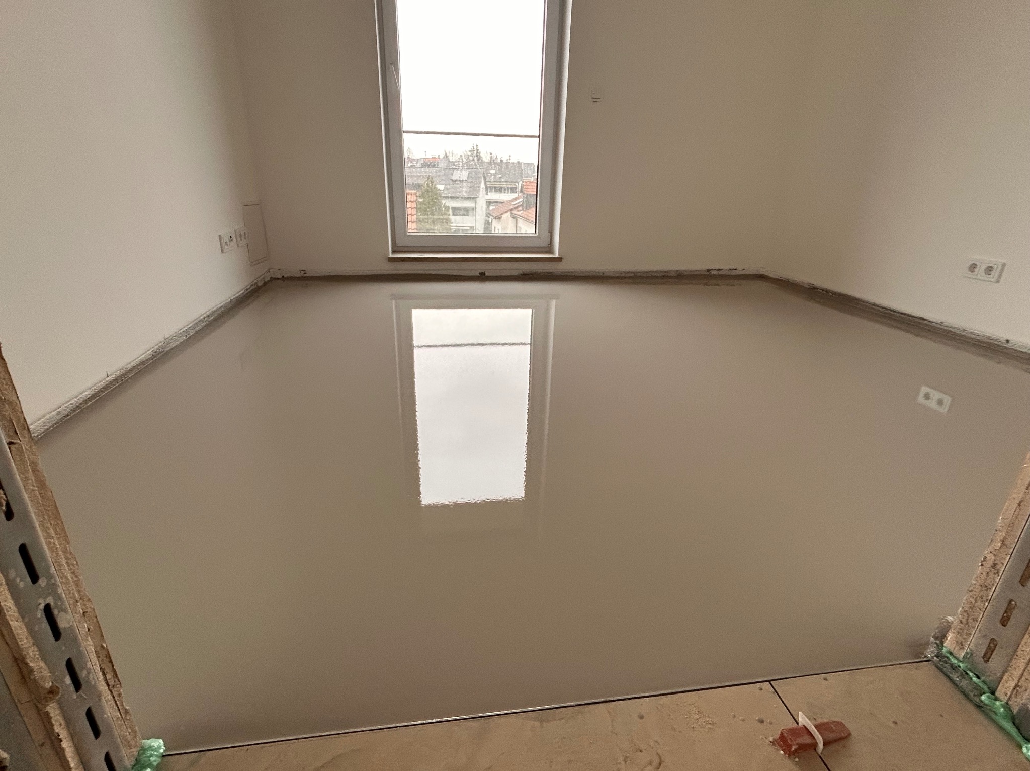 Świeżo wylana, gładka posadzka anhydrytowa w jasnym pomieszczeniu z oknem, odbijającym światło. Widoczne listwy przypodłogowe i fragment rusztowania.