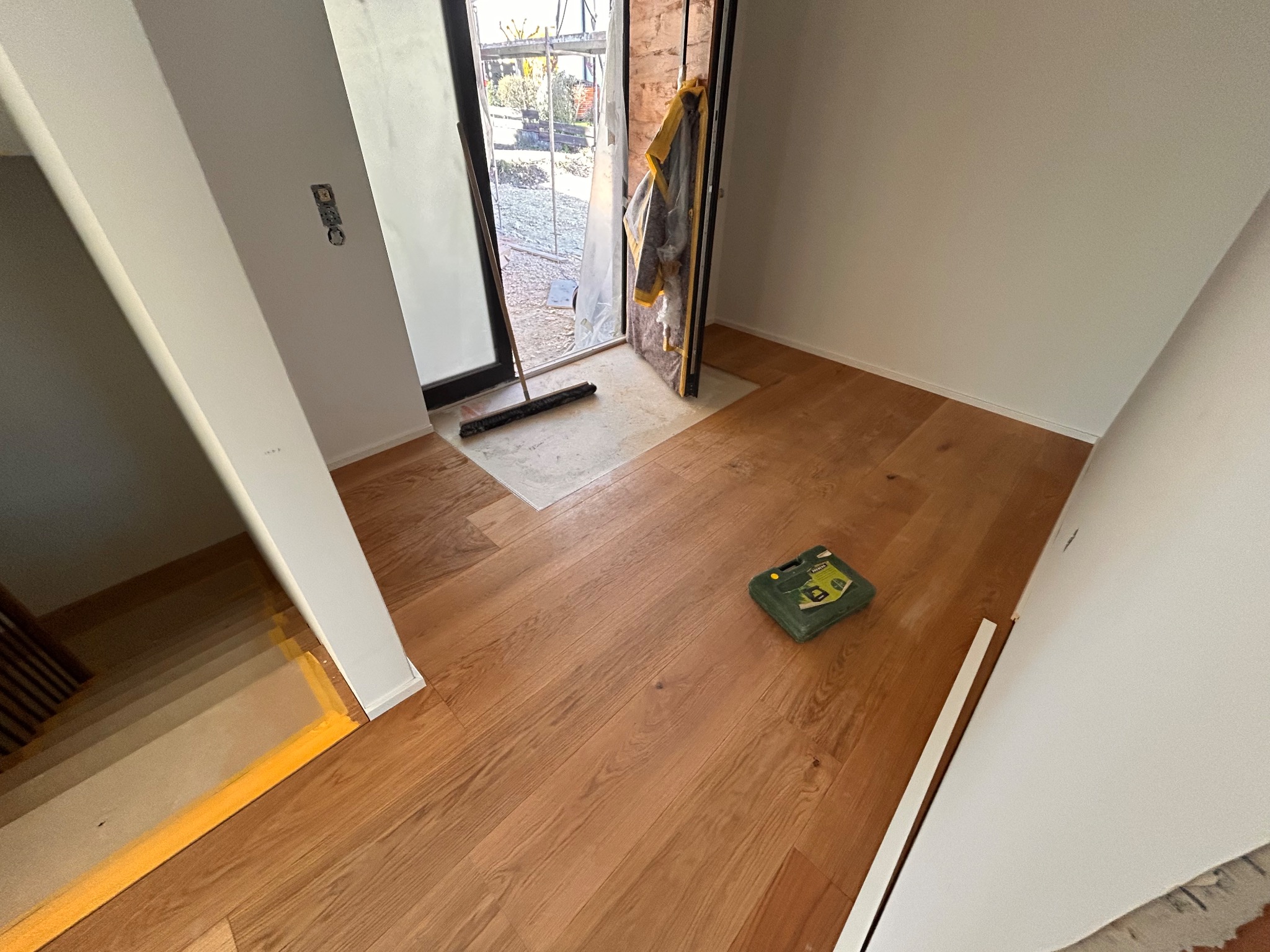 Jasny parkiet w nowym domu, widok na schody i otwarte drzwi z narzędziami. Zielona skrzynka narzędziowa leży na podłodze. Widoczne listwy przypodłogowe.