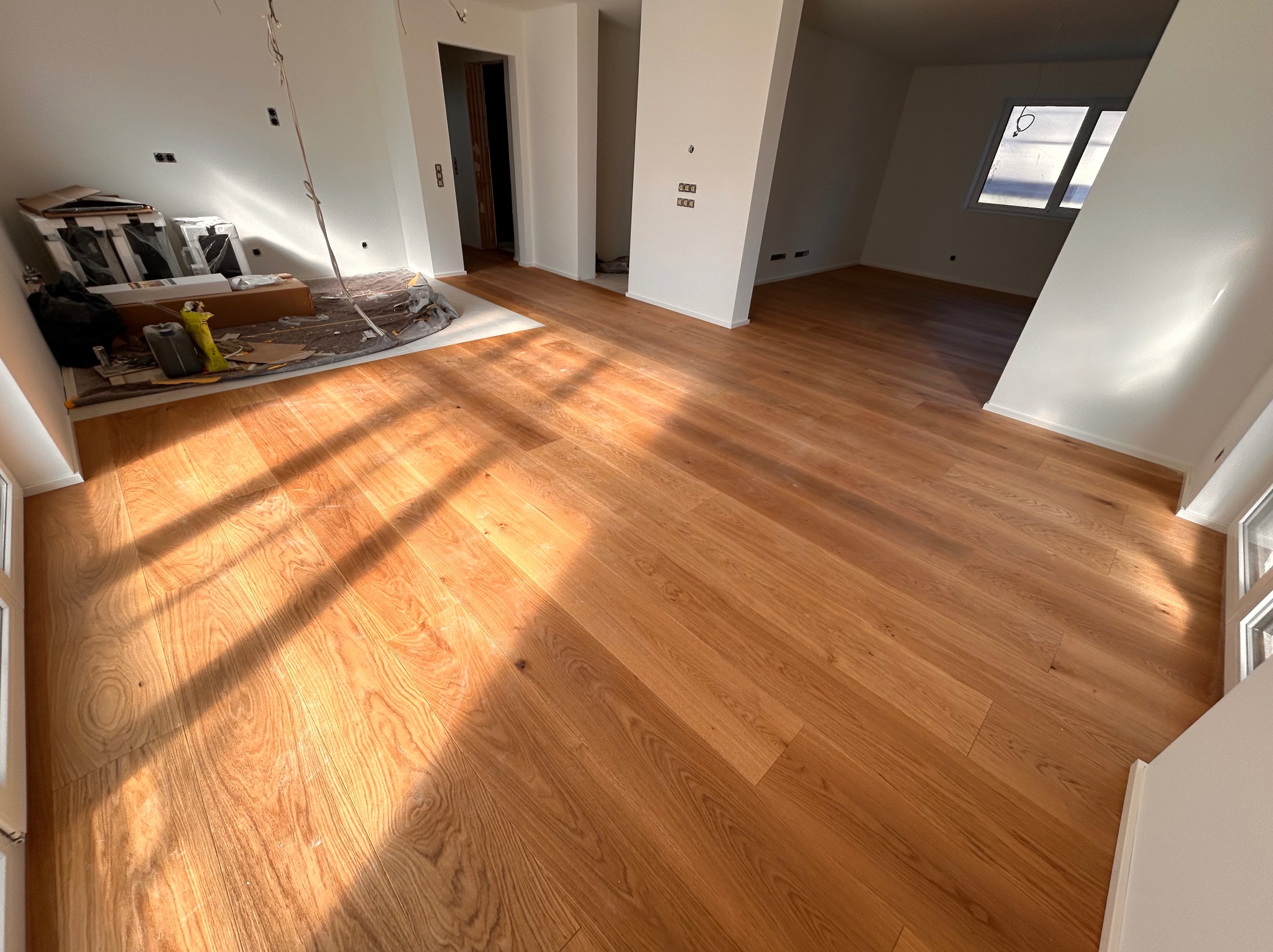 Jasny parkiet w nowym domu, oświetlony promieniami słońca. Widoczne materiały budowlane w rogu pokoju, podkreślające świeżość wykończenia.