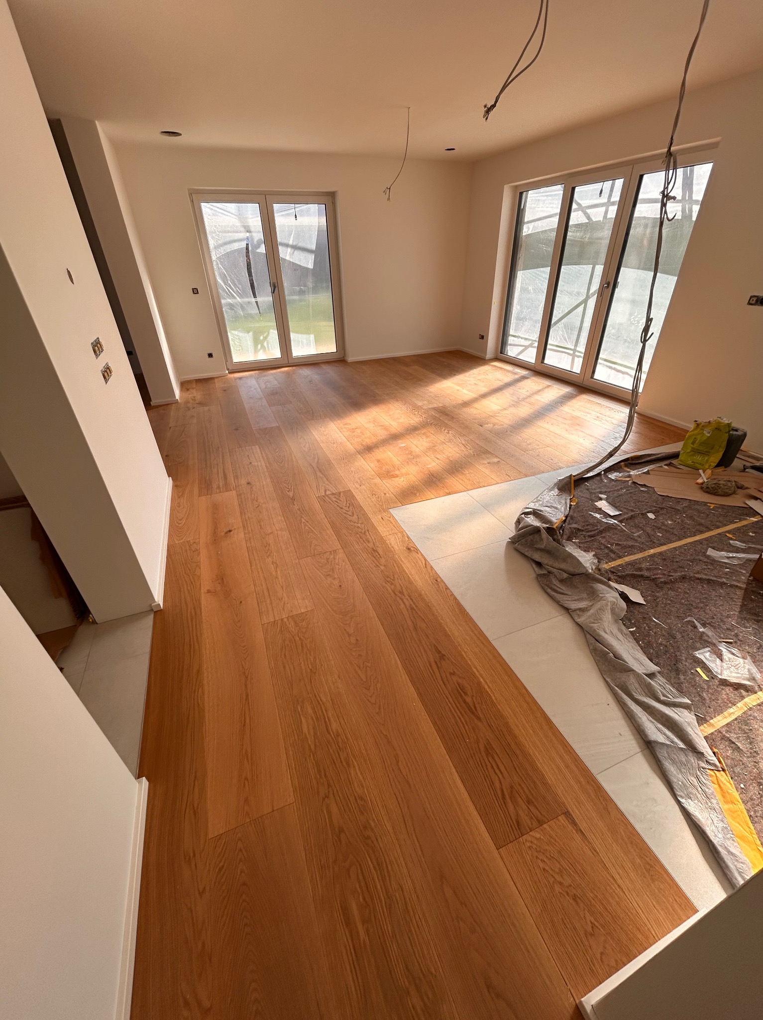 Częściowo ułożony parkiet w jasnym pokoju z dużymi oknami. Widoczne zabezpieczenia podłogi i wystające kable. Naturalne światło podkreśla strukturę drewna.