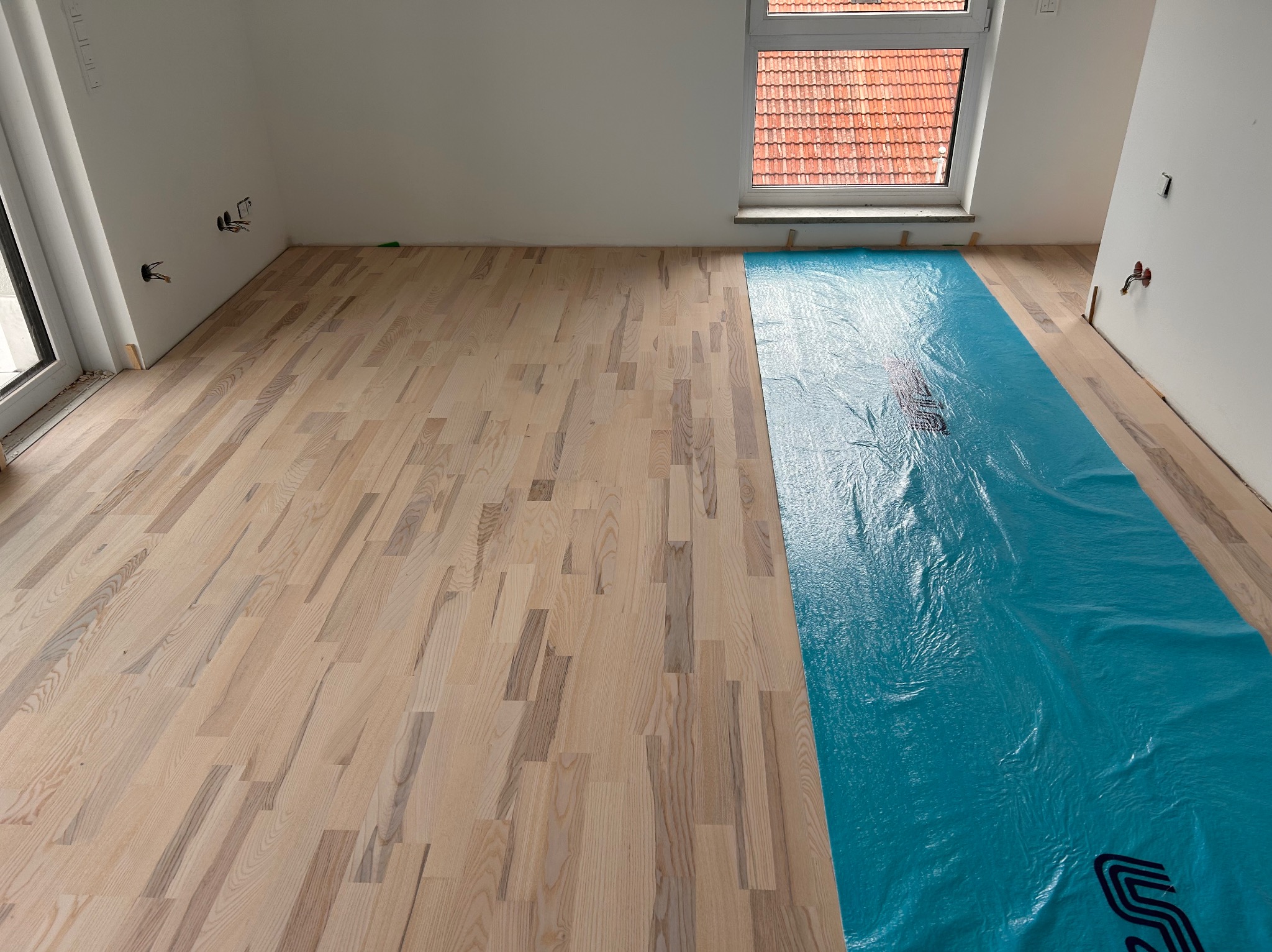 Jasny parkiet ułożony w nowym wnętrzu, częściowo zabezpieczony niebieską folią ochronną. Widoczne gniazdka elektryczne i okno z widokiem na dachówkę.