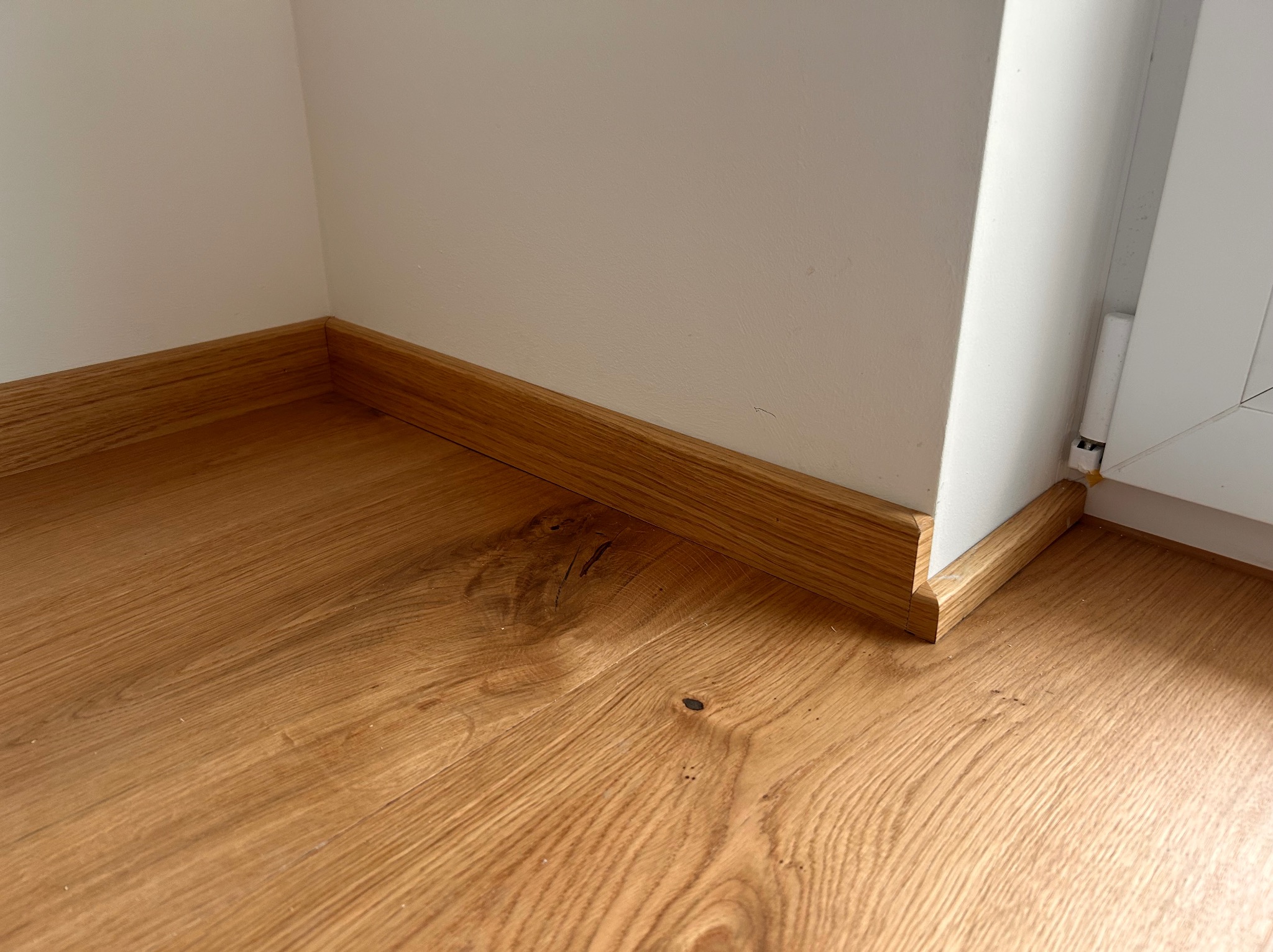 Jasny parkiet z drewnianą listwą przypodłogową przy białej ścianie i futrynie drzwi. Widok z niskiego kąta, ukazujący detale wykończenia podłogi.