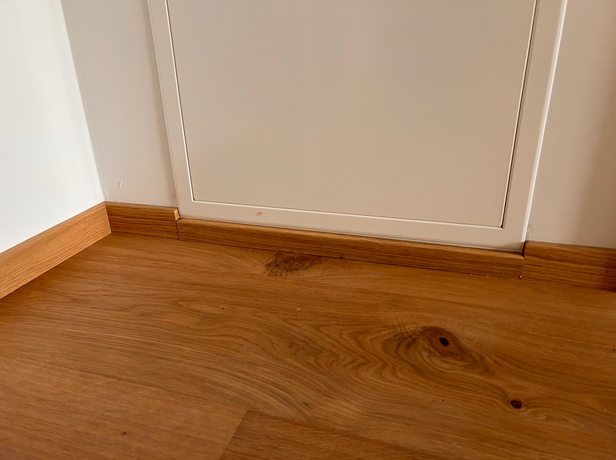 Jasny parkiet z drewnianą listwą przypodłogową i białymi drzwiczkami rewizyjnymi. Widoczne słoje drewna i detale wykończenia.