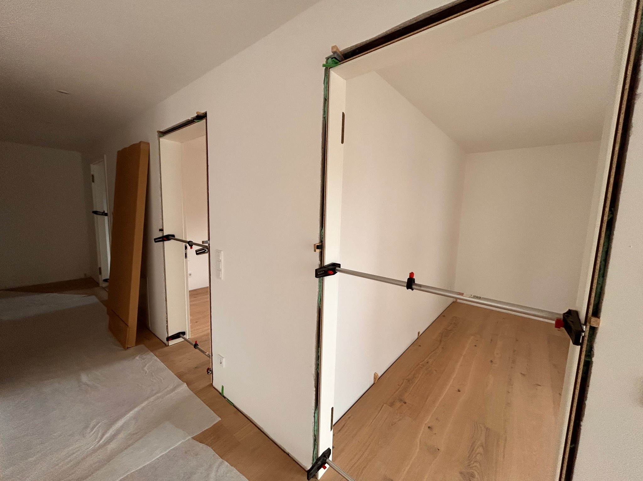 Wnętrze z zamontowanymi ościeżnicami drzwiowymi, zabezpieczonymi rozpórkami. Na podłodze widoczny parkiet, częściowo zakryty folią ochronną. Ściany pomalowane na biało.