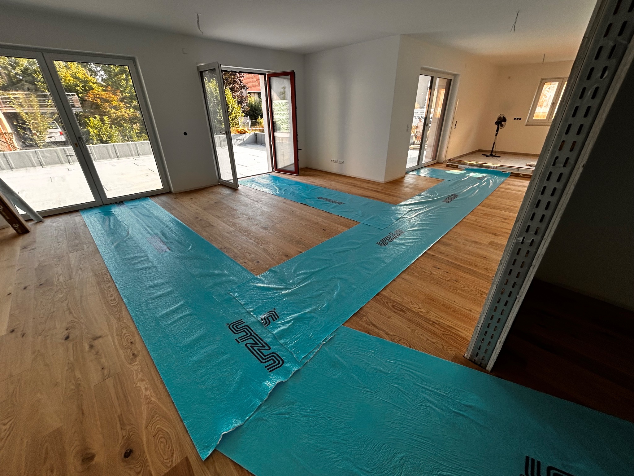 Jasne drewniane deski podłogowe częściowo zakryte turkusową folią ochronną w przestronnym, jasnym wnętrzu z dużymi oknami balkonowymi i otwartymi drzwiami.