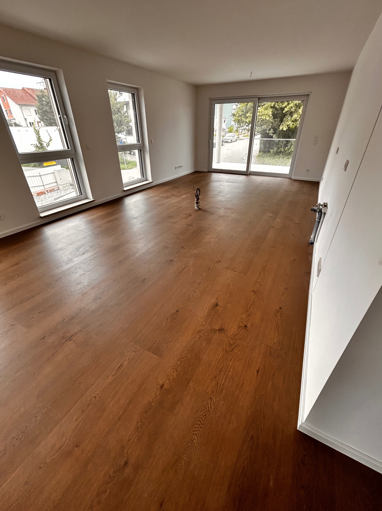 Salon z nową, brązową podłogą z paneli, widok na okna i drzwi balkonowe. Minimalistyczne wnętrze z białymi ścianami i jasnym światłem dziennym.