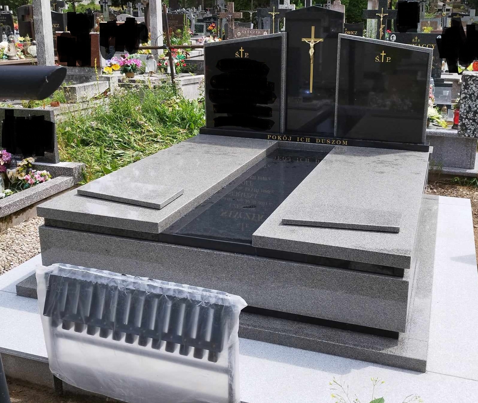 Nowoczesny pomnik z ciemnego granitu z wygrawerowanym krzyżem i napisem 'Pokój Ich Duszom' na cmentarzu, widoczne inne nagrobki w tle, w dolnej części zdjęcia osłona na znicze.