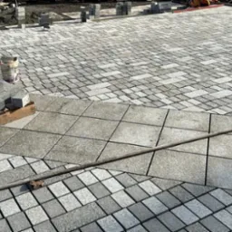 Układanie kostki granitowej: widok na fragment powierzchni z ułożoną kostką granitową w trzech różnych wzorach, obok stos kostki przygotowanej do ułożenia i narzędzia.