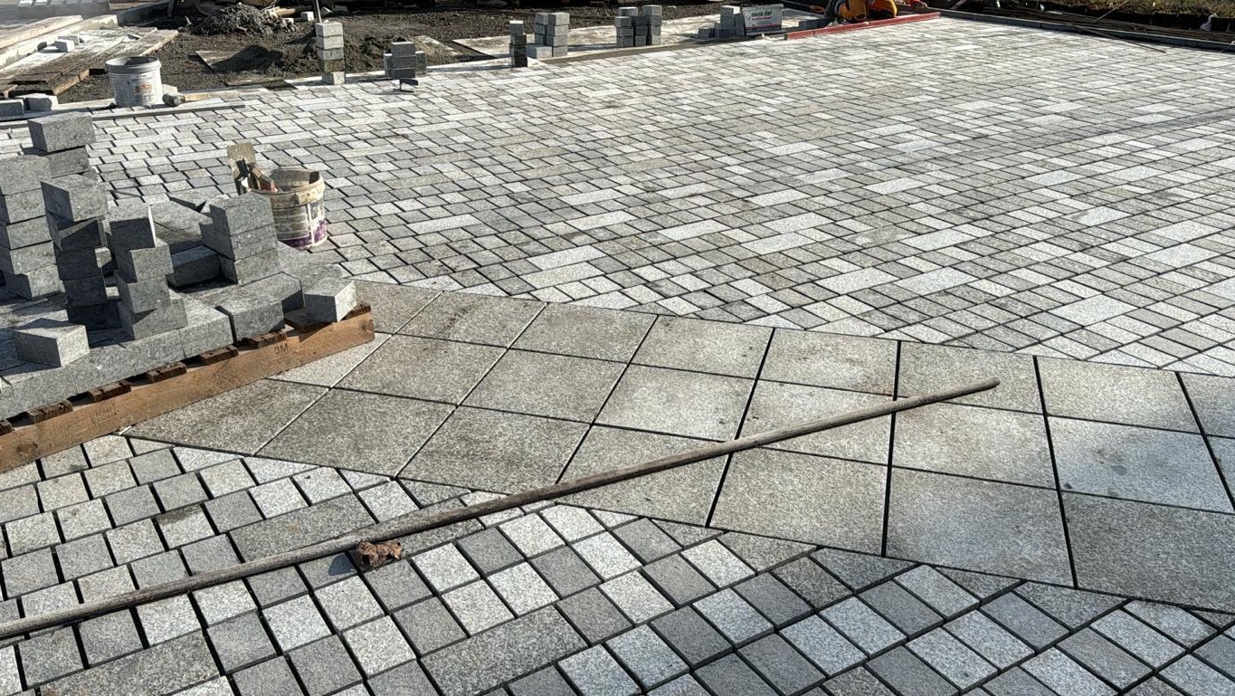 Układanie kostki granitowej: widok na fragment powierzchni z ułożoną kostką granitową w trzech różnych wzorach, obok stos kostki przygotowanej do ułożenia i narzędzia.