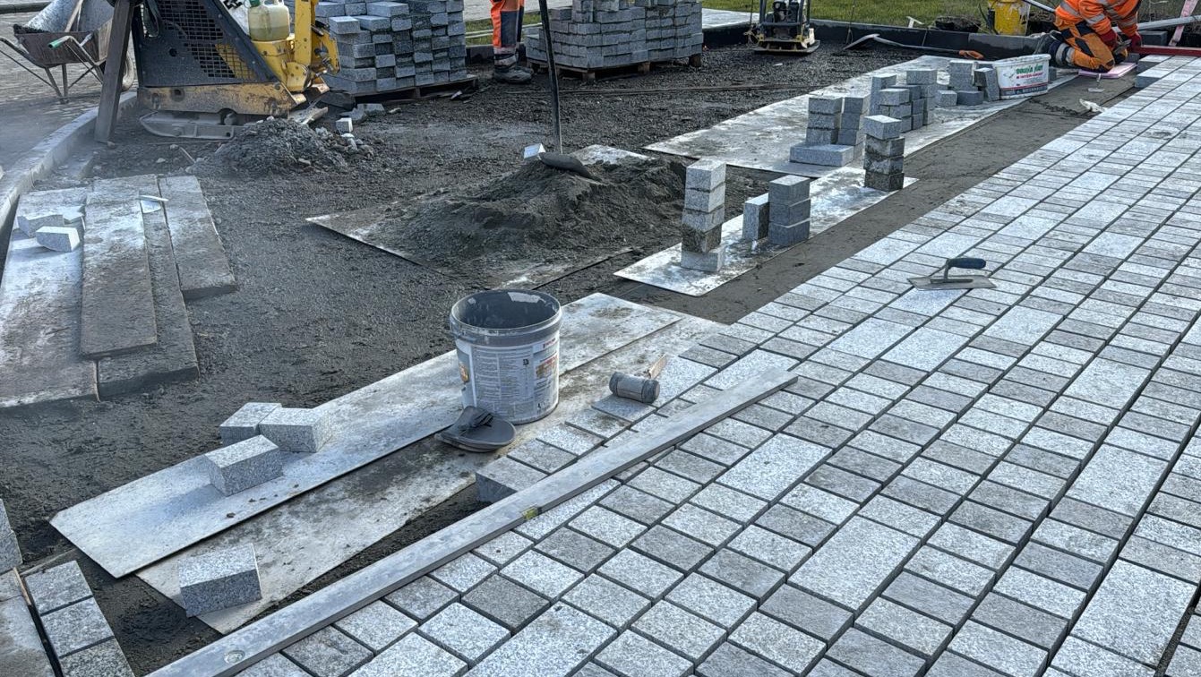 Szara kostka granitowa układana na podsypce piaskowej, widoczny poziomica, gumowy młotek, wiadro z zaprawą, ubijarka, oraz paleta z kostką w tle.