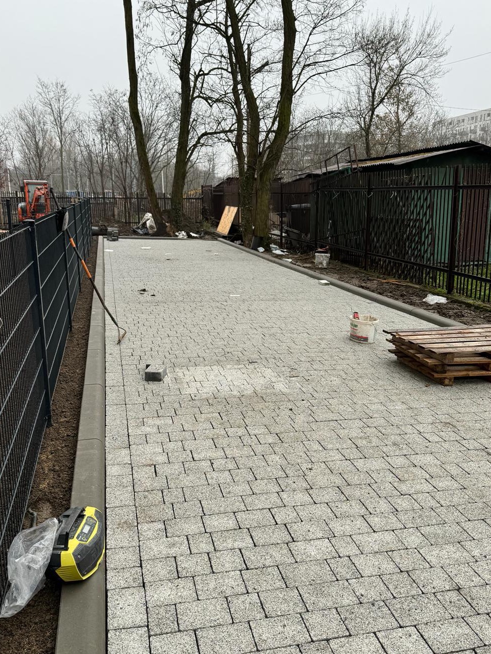 Ułożona szara kostka brukowa na posesji z widocznym sprzętem budowlanym, paletami i narzędziami, w tle drzewa i budynki gospodarcze.