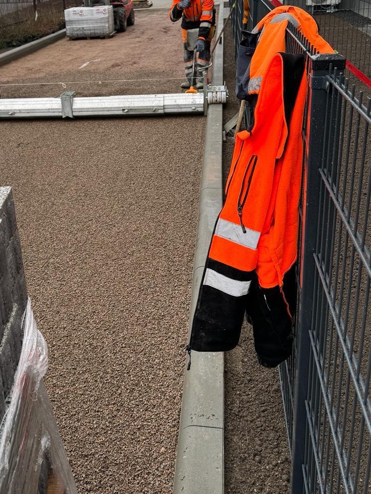 Przygotowanie podłoża pod kostkę brukową: zagęszczona warstwa podsypki, widoczna maszyna do zagęszczania i paleta kostki w tle, pomarańczowa kurtka robocza na ogrodzeniu.