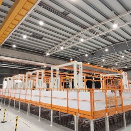 Joka Industry Joanna Kasperska - Wnętrze hali produkcyjnej z konstrukcją metalową pomalowaną na biało-pomarańczowo, prawdopodobnie etap montażu, widoczny pracownik na drabinie, suwnica pod sufitem.