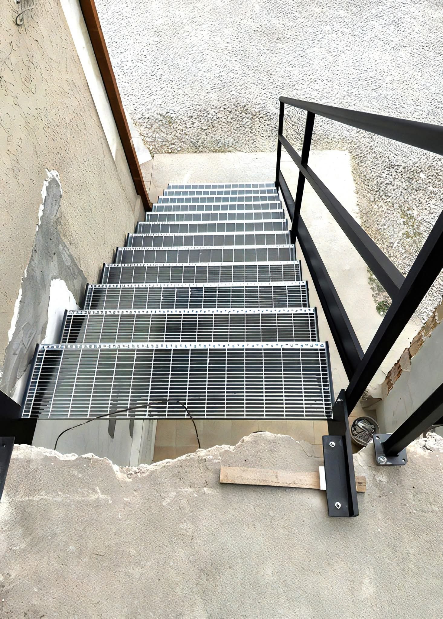 Metalowe schody kratowe z czarną balustradą, widok z góry na betonowe podłoże w trakcie montażu, z widocznymi elementami mocującymi i deską stabilizującą.