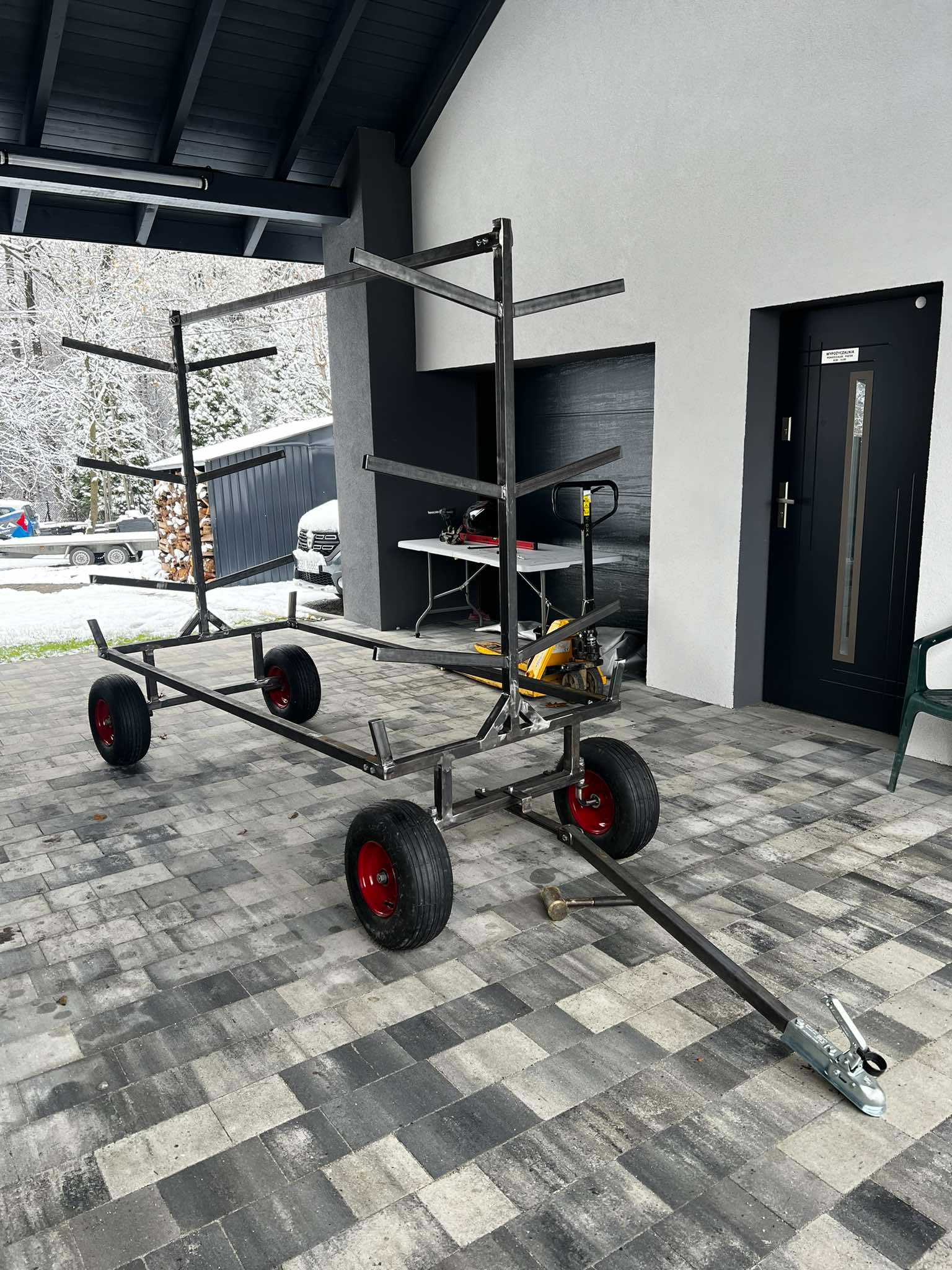 Wózek transportowy z metalową konstrukcją regałową na czterech kołach z czerwoną felgą, z dyszlem zakończonym zaczepem kulowym, stojący na brukowanym podjeździe przed domem z czarnymi elementami...