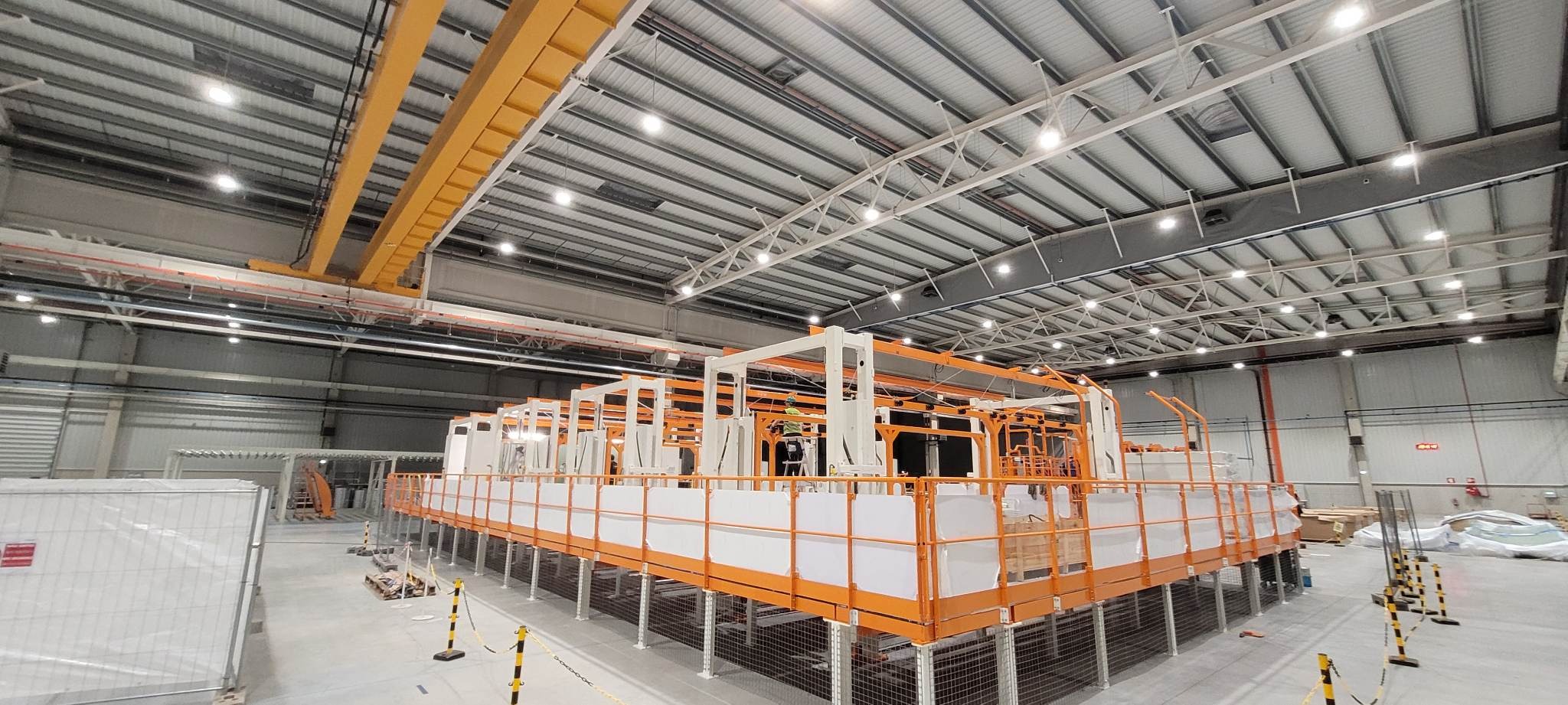 Wnętrze hali produkcyjnej z konstrukcją metalową pomalowaną na biało-pomarańczowo, prawdopodobnie etap montażu, widoczny pracownik na drabinie, suwnica pod sufitem.