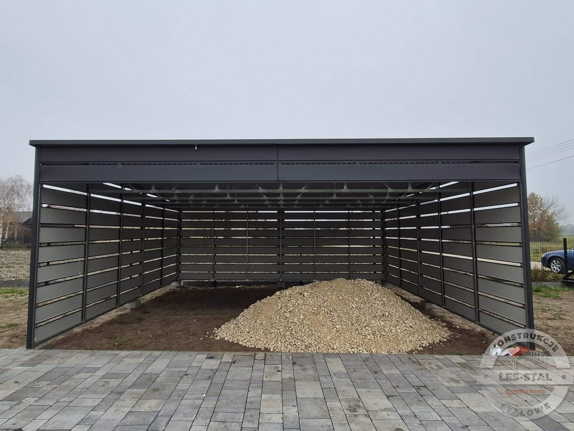 Nowoczesna pergola metalowa w kolorze grafitowym z poziomymi panelami, wypełniona stertą żwiru na posesji z kostki brukowej w pochmurny dzień.