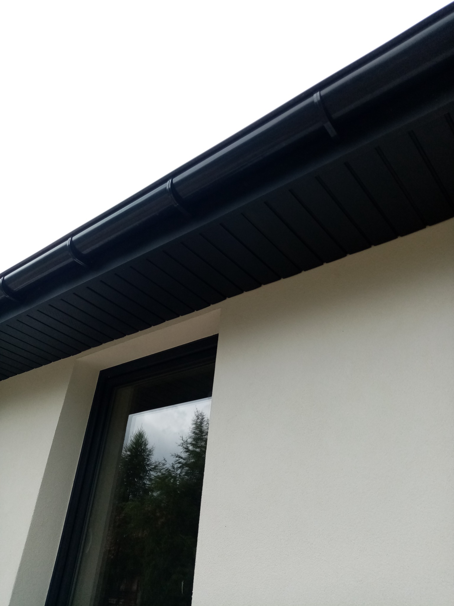 Elewacja budynku z oknem i czarnym orynnowaniem. Widok z dołu, podkreślający nowoczesny design i precyzję wykonania detali.