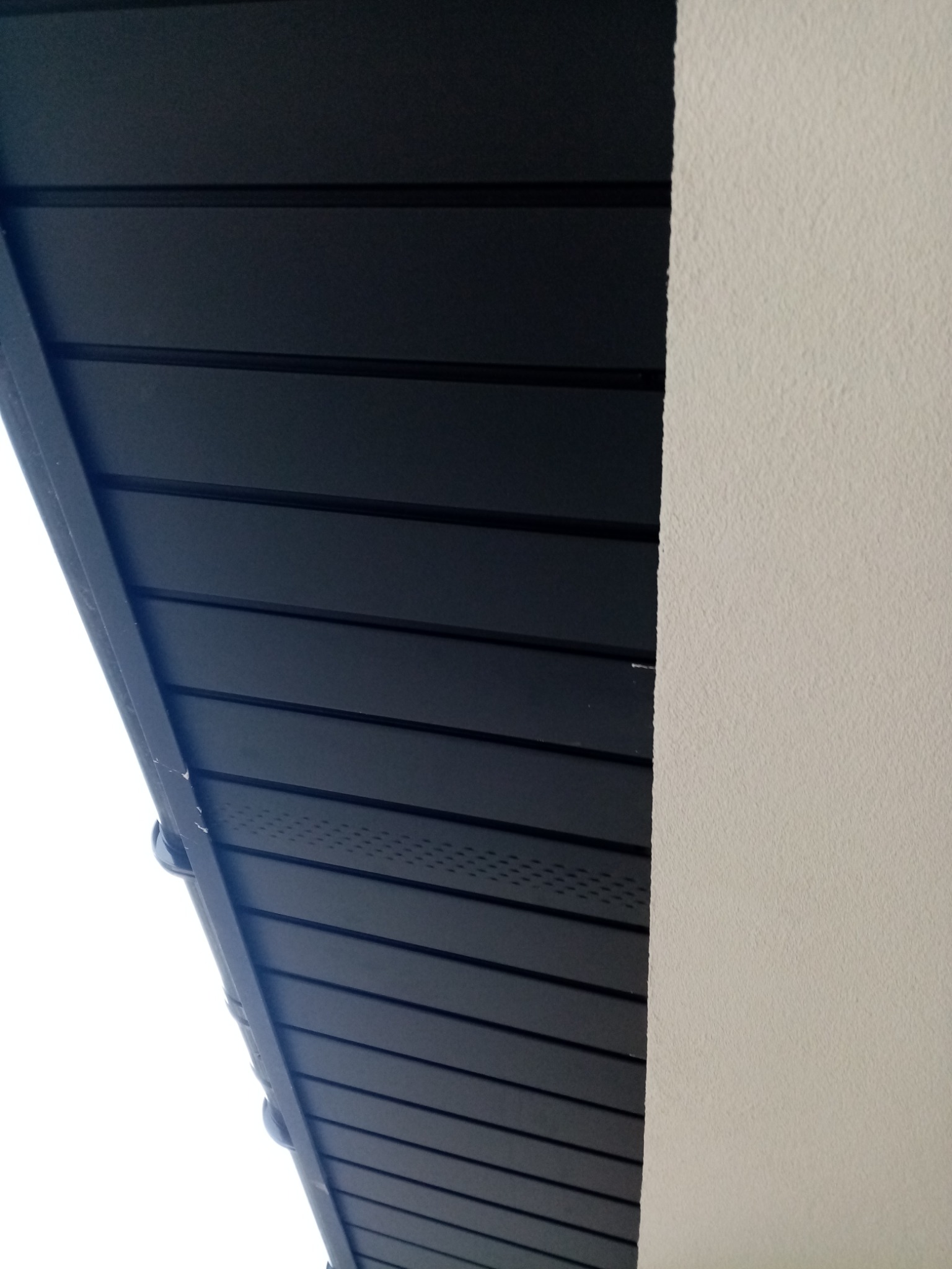 Fragment elewacji budynku z ciemną, poziomą okładziną i jasną, teksturowaną ścianą. Widok z dołu, podkreślający kontrast materiałów i kolorów.