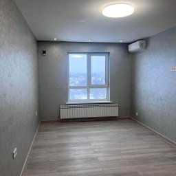 Jurii Kostaszczuk - Pusty pokój po remoncie z szarymi ścianami, podłogą imitującą drewno, białym grzejnikiem pod oknem i lampą sufitową.