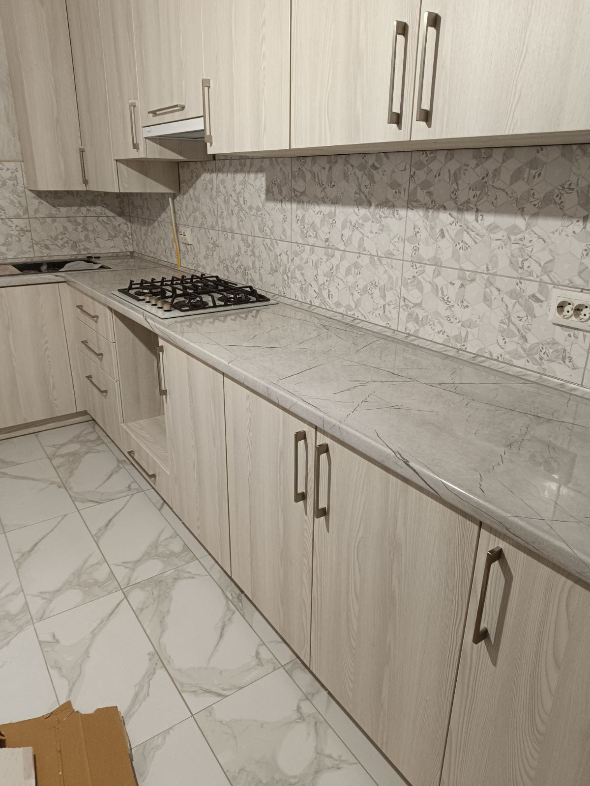 Jasna kuchnia z drewnopodobnymi szafkami, marmurowym blatem i wzorzystymi płytkami na ścianie.