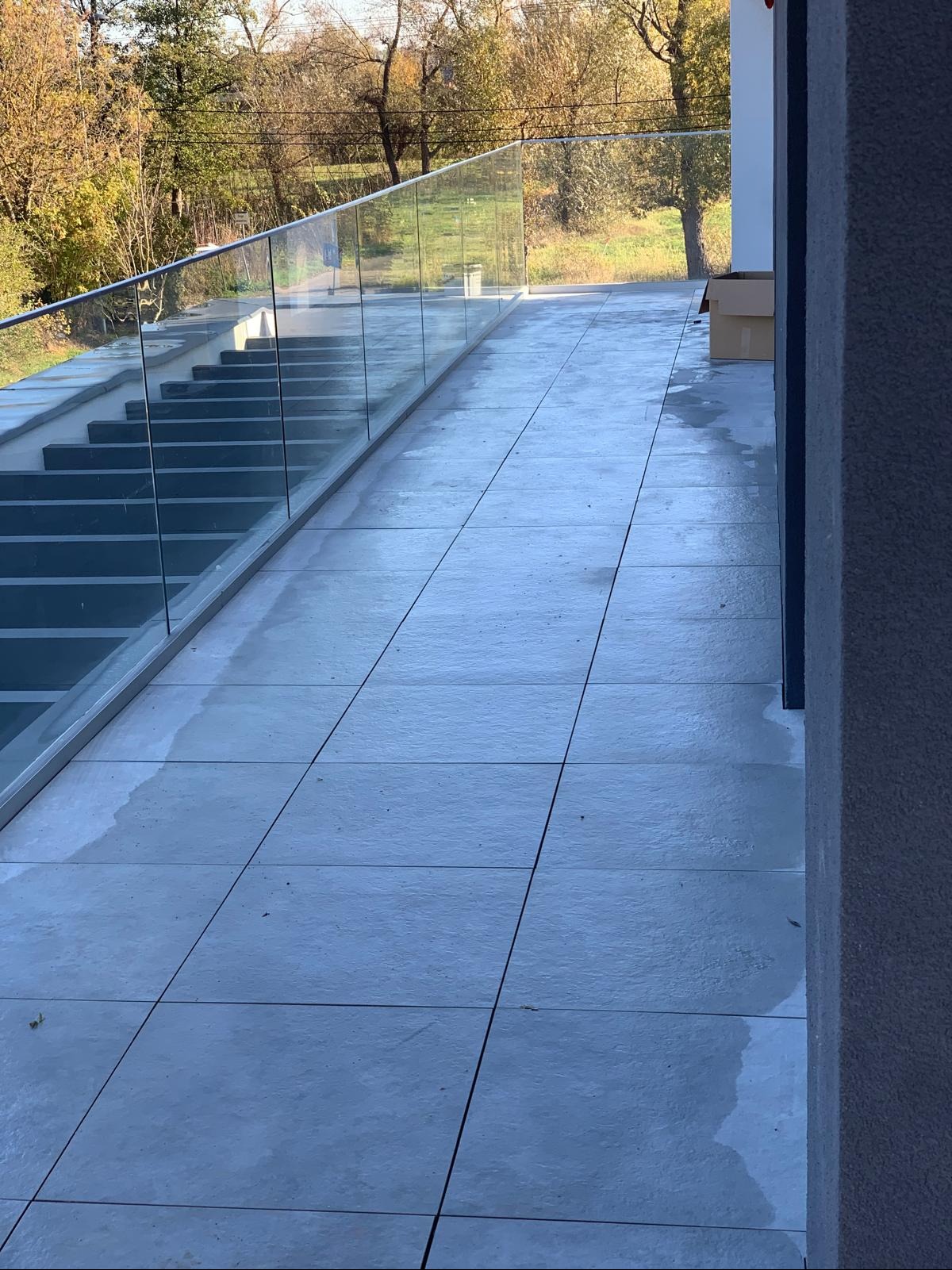 Szare płytki ułożone na zewnątrz budynku, widoczne szklane balustrady i schody, w tle drzewa.