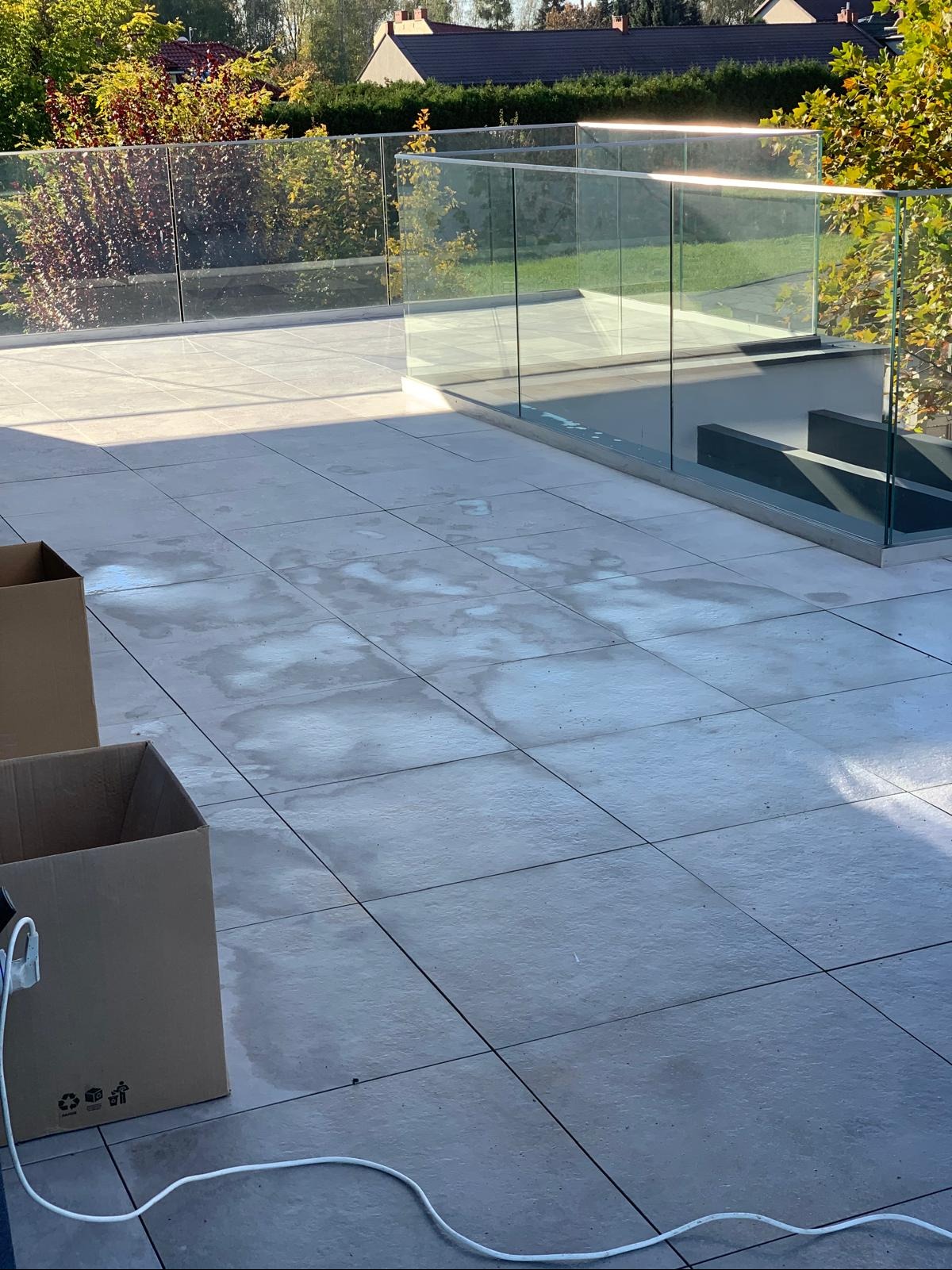 Szare płytki tarasowe z widocznymi mokrymi plamami, szklana balustrada z metalowymi elementami, kartony i kabel zasilający w rogu, widok na zieleń i zabudowę w tle.
