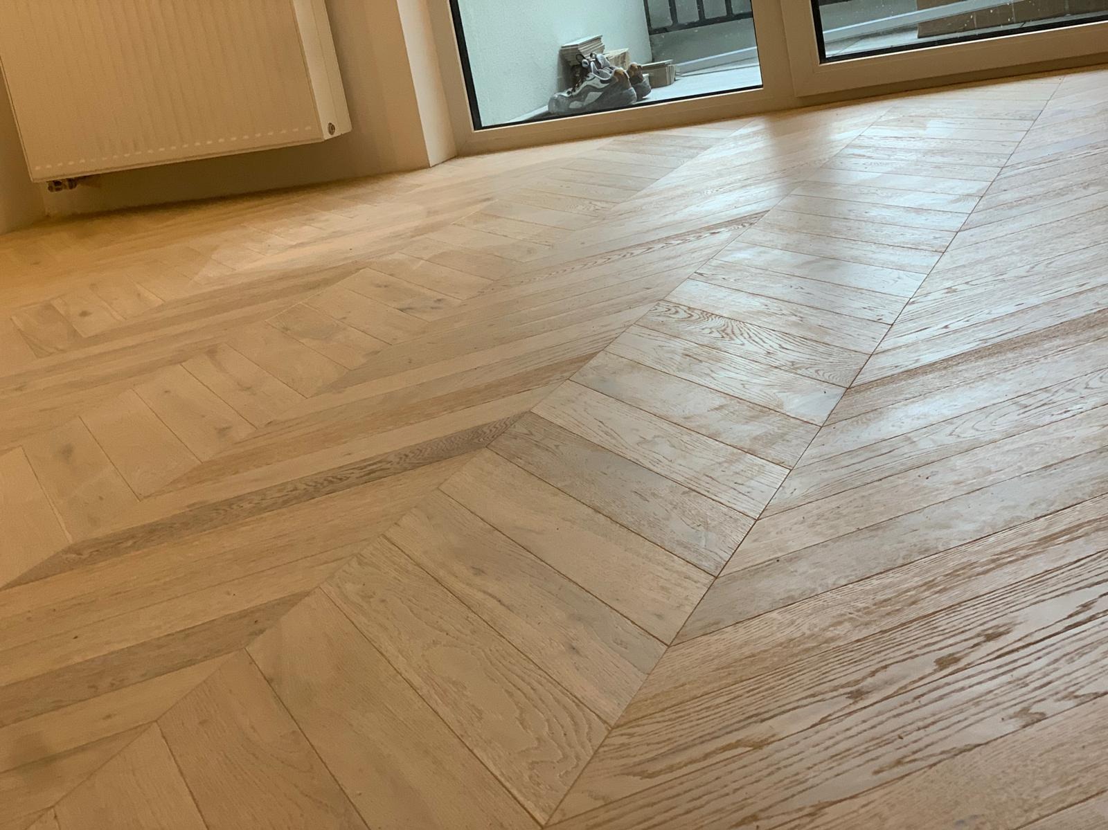 Jasna, drewniana podłoga ułożona w jodełkę francuską, widok na okno z butami na parapecie i grzejnik na ścianie.