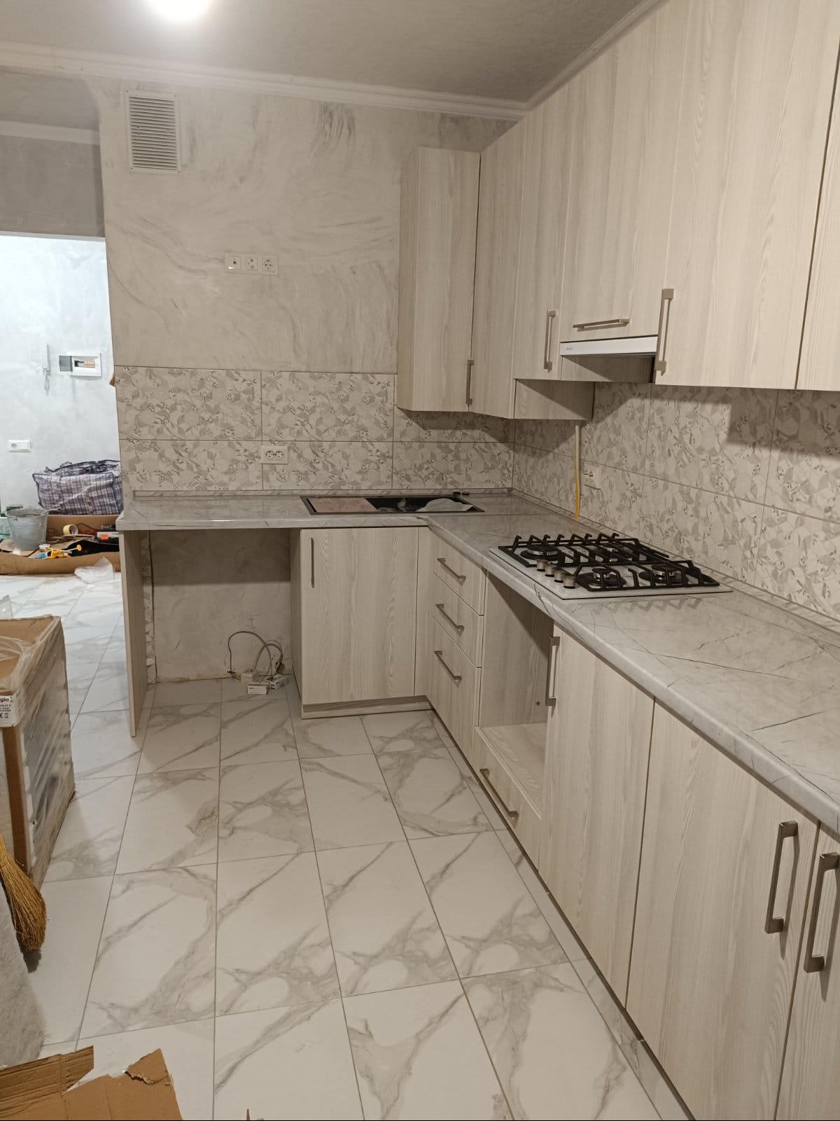 Jasna kuchnia z marmurowymi płytkami na podłodze i ścianie nad blatem, widoczna płyta gazowa, szafki z drewnopodobnym wykończeniem, w tle pomieszczenie w trakcie prac wykończeniowych.