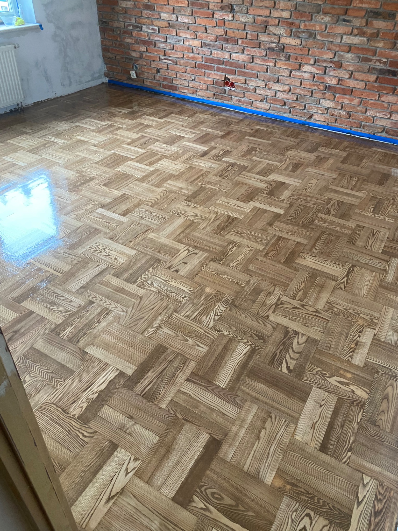 Nowo ułożony parkiet w jodełkę francuską, z widocznym połyskiem świeżego lakieru, zabezpieczony niebieską taśmą malarską przy ceglanej ścianie.