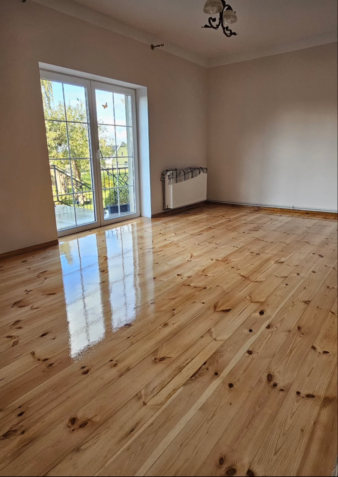 Świeżo polakierowana, błyszcząca podłoga z desek w pustym pokoju, odbijająca światło z okna balkonowego.