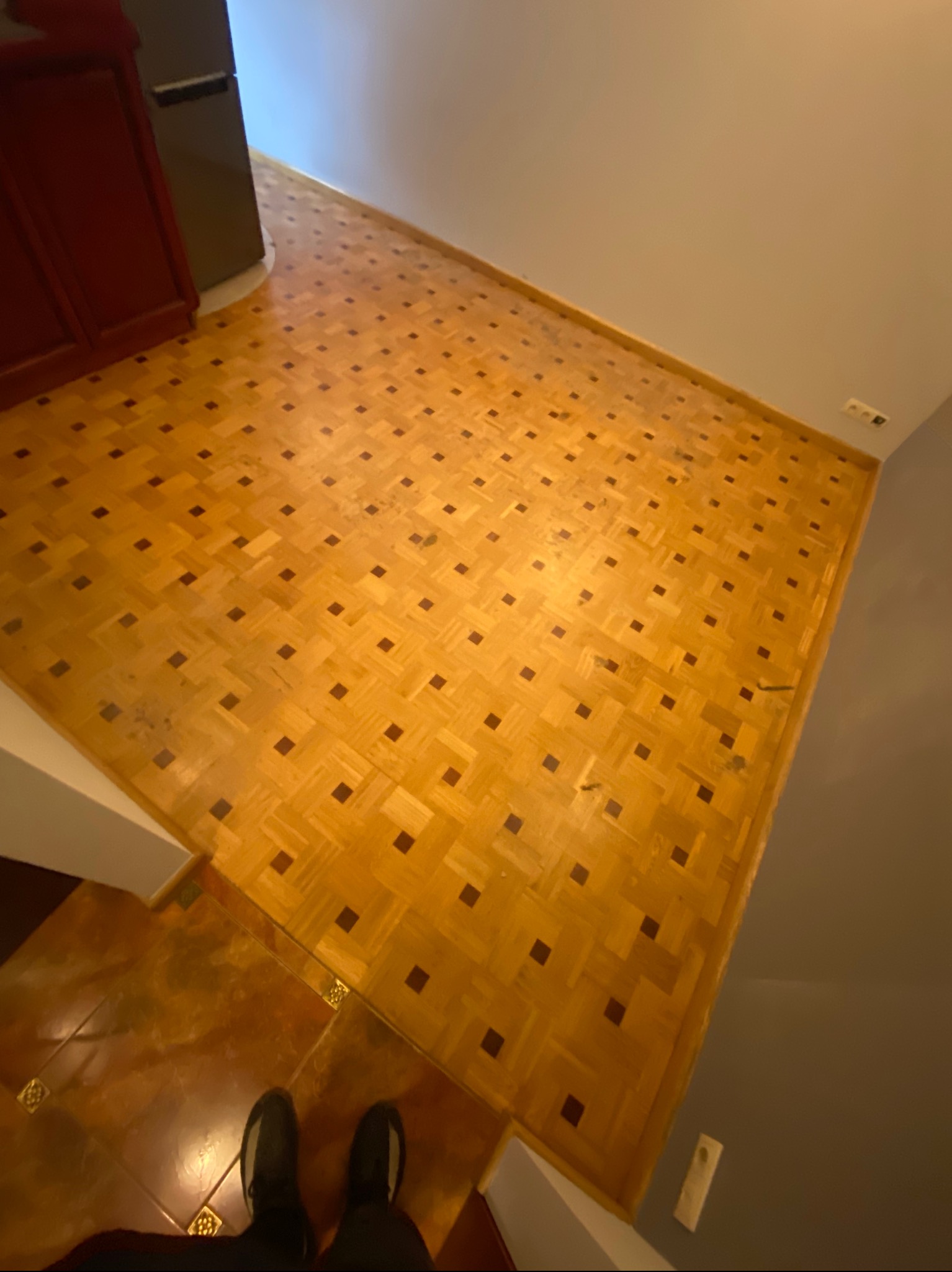 Parkiet z jasnego drewna ułożony w kwadraty z ciemniejszymi wstawkami, widoczne ślady użytkowania i dwie osoby w czarnych butach w dolnej części kadru.