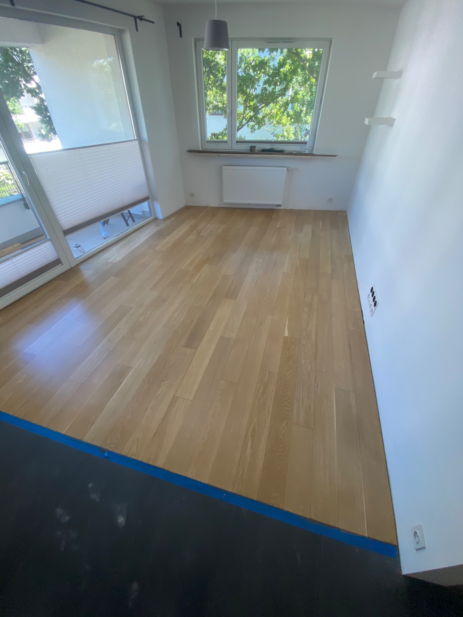 Jasny, świeżo wycyklinowany parkiet w pokoju z białymi ścianami, widoczne okno z roletami i fragment niebieskiej taśmy malarskiej na podłodze w rogu pomieszczenia.