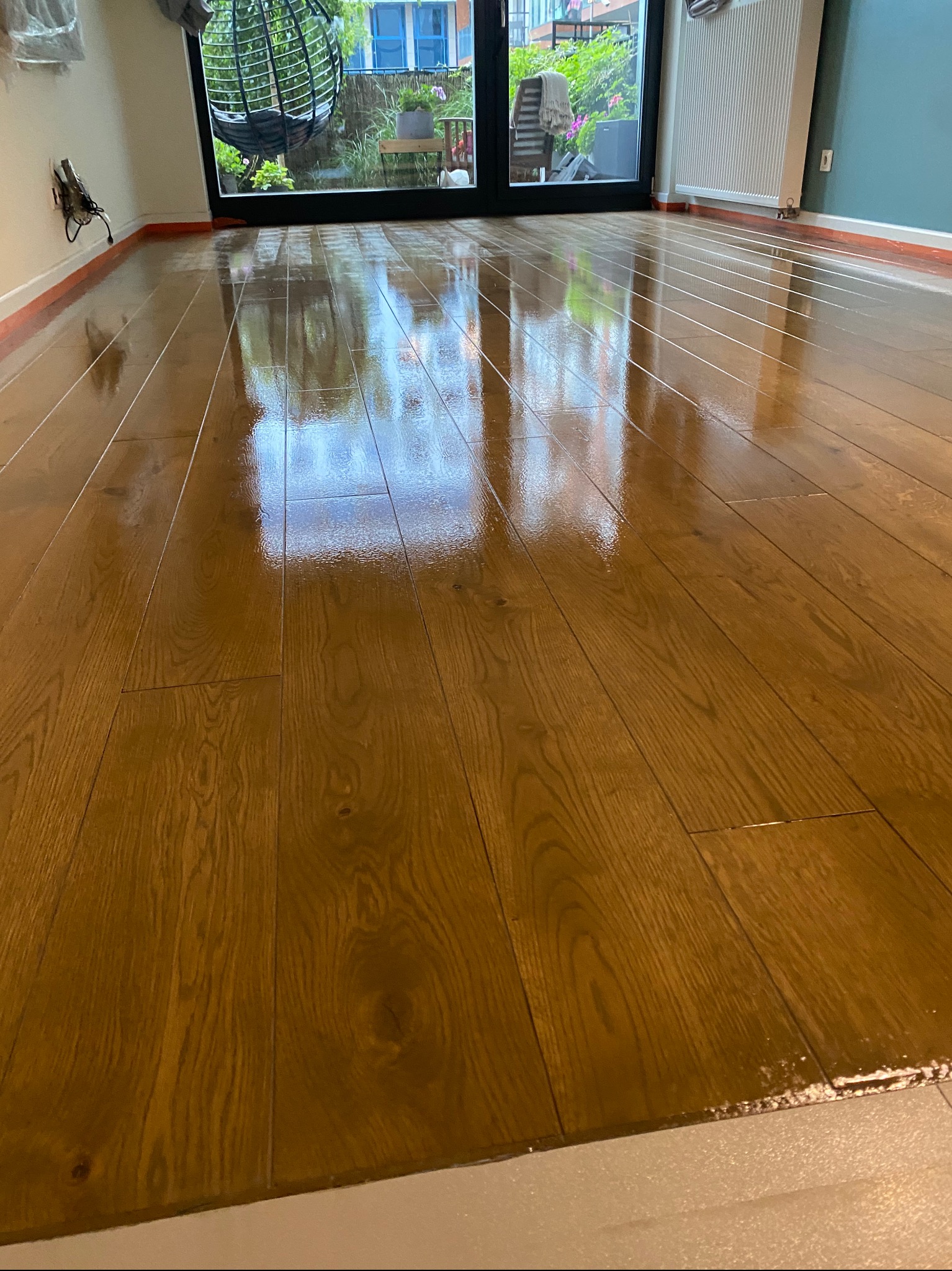 Świeżo polakierowana, błyszcząca podłoga z drewnianych desek, z widocznym odbiciem okna i elementów wystroju wnętrza, wykończona listwą przypodłogową. Na podłodze widoczne drobne niedoskonałości...