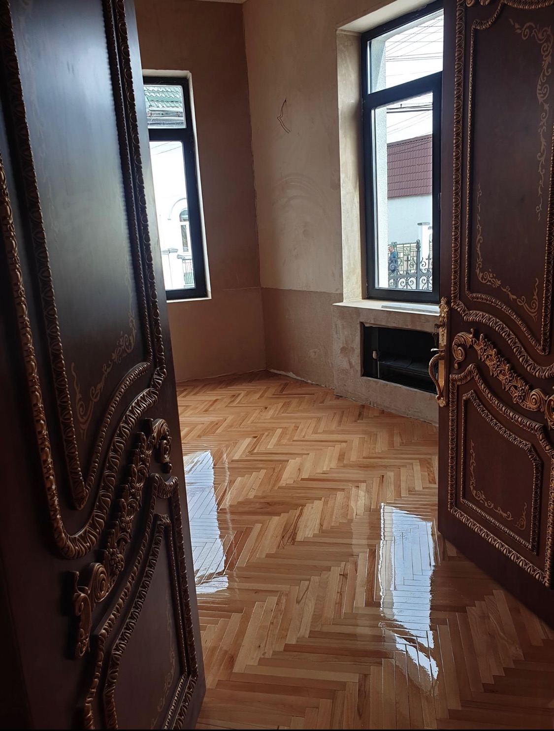 Świeżo położony parkiet w jodełkę, odbijający światło w przestronnym pokoju z otwartymi, bogato zdobionymi drzwiami i widokiem na zewnątrz przez okna.