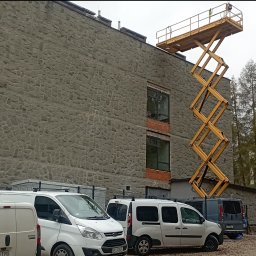 R1 Trade Dmytro Rupchev - Żółty podnośnik koszowy ustawiony przy budynku z kamienną elewacją, widoczne okna z ceglanym obramowaniem i zaparkowane busy.