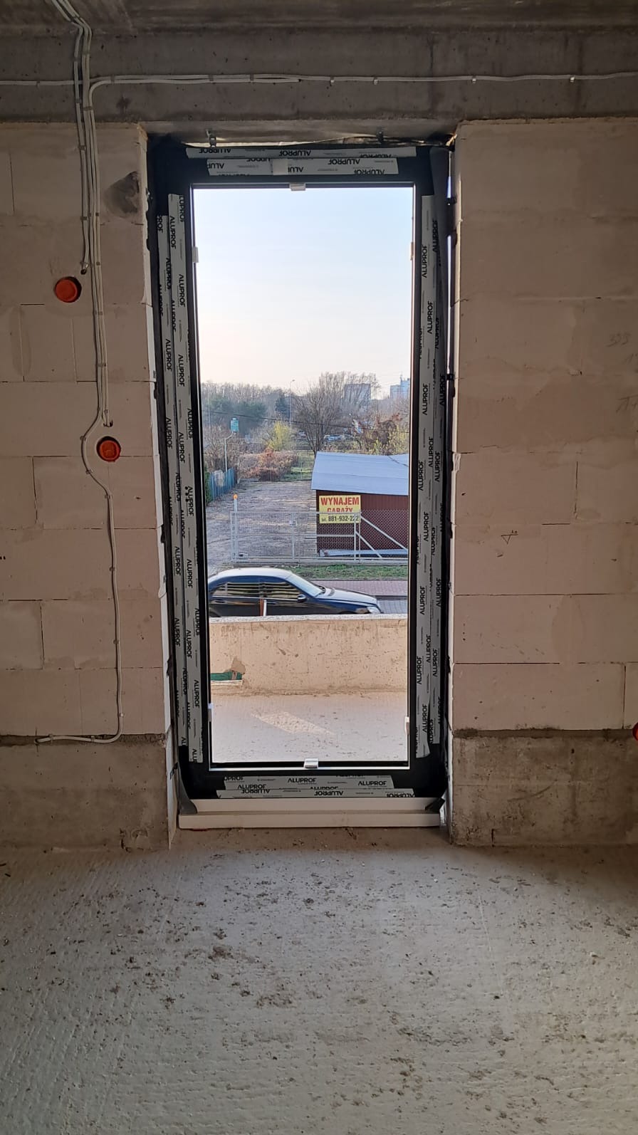 Montaż okna aluminiowego w surowym wnętrzu, widoczne przewody elektryczne i budynek z napisem 'Wynajem' w tle.