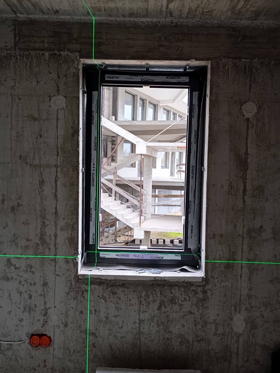 Montaż okna w surowym wnętrzu, widoczne ściany betonowe, laserowe linie poziomowania, okno z czarną ramą z widokiem na schody i budynek w trakcie budowy.